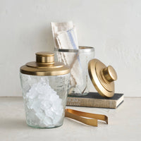 Decorative glass ice buckets with tongs and hammered gold lids on a light surface.