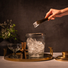 Hand using tongs to grab ice from a pebbled glass ice bucket on a gold tray with a dark background.