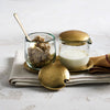 Small pebbled glass jars with hammered gold lids and spoons on a textured surface