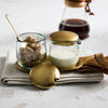 Two small pebbled glass jars with hammered gold lids, one containing sugar and the other cream, with a coffee carafe in the background.